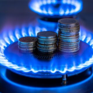 Stosy monet ułożone na palniku gazowym z niebieskim płomieniem, symbolizujące wysokie koszty zużycia gazu i opłat za energię.