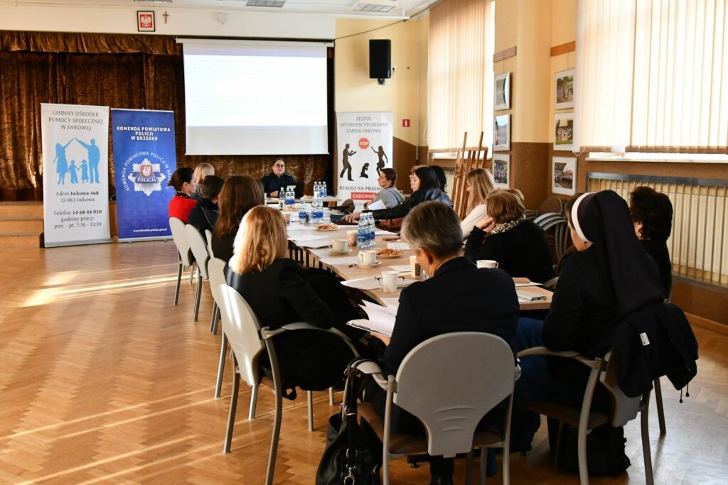 Zdjęcie przedstawia uczestników siedzących przy dużym, prostokątnym stole w jasnej sali konferencyjnej. Na stole leżą dokumenty, napoje i przekąski. W tle widać projektor z prezentacją oraz trzy stojące roll-upy instytucji zajmujących się pomocą społeczną i przeciwdziałaniem przemocy. Osoby biorące udział w spotkaniu słuchają prelegenta siedzącego na końcu stołu