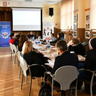 Uczestnicy siedzą wokół dużego stołu w jasnej sali podczas spotkania lub szkolenia. Na stole stoją butelki z wodą, filiżanki i dokumenty. W tle widoczne są trzy roll-upy instytucji oraz ekran z prezentacją. Osoby słuchają prelegenta siedzącego na końcu stołu.