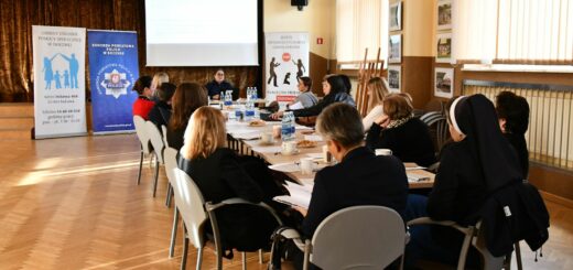 Uczestnicy siedzą wokół dużego stołu w jasnej sali podczas spotkania lub szkolenia. Na stole stoją butelki z wodą, filiżanki i dokumenty. W tle widoczne są trzy roll-upy instytucji oraz ekran z prezentacją. Osoby słuchają prelegenta siedzącego na końcu stołu.