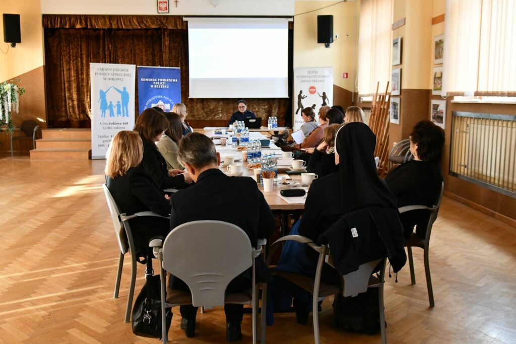 Uczestnicy siedzą wokół dużego stołu w jasnej sali podczas spotkania lub szkolenia. Na stole stoją butelki z wodą, filiżanki i dokumenty. W tle widoczne są trzy roll-upy instytucji oraz ekran z prezentacją. Osoby słuchają prelegenta siedzącego na końcu stołu