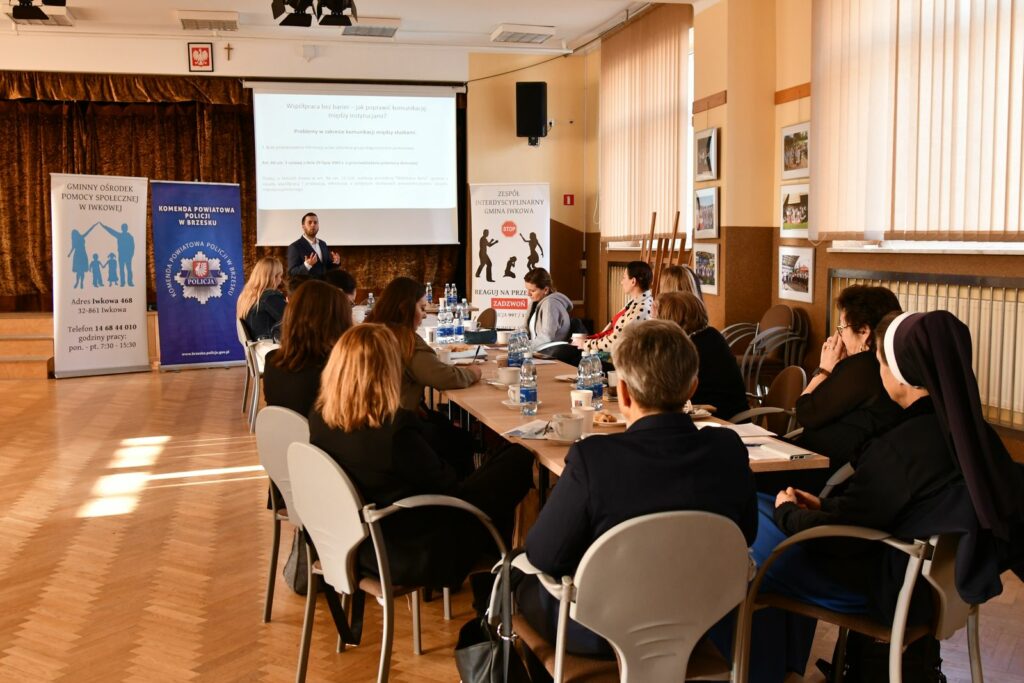 Prelegent stoi przed ekranem z wyświetloną prezentacją i przemawia do uczestników siedzących przy dużym stole w sali konferencyjnej. Na stole stoją butelki z wodą, filiżanki i dokumenty. W tle widoczne są trzy roll-upy instytucji zajmujących się pomocą społeczną, policją oraz przeciwdziałaniem przemocy.