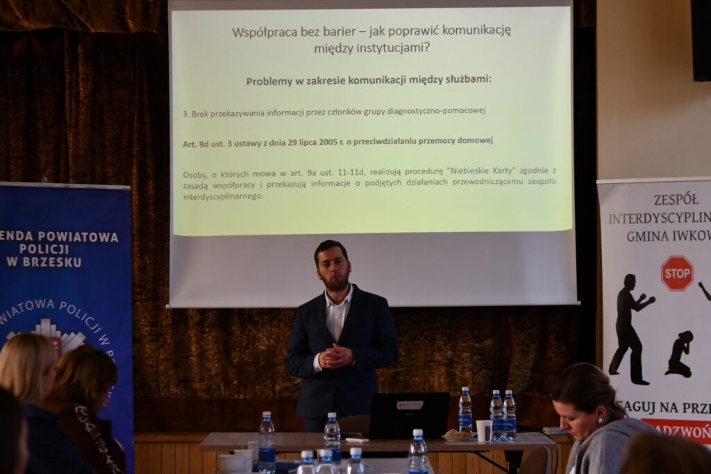 Prelegent w garniturze stoi przed ekranem z wyświetloną prezentacją dotyczącą współpracy między instytucjami i komunikacji między służbami. Przed nim na stole stoją butelki z wodą i laptop. Po bokach widoczne są roll-upy policji oraz Zespołu Interdyscyplinarnego w Iwkowej, a na pierwszym planie siedzą uczestnicy spotkania.