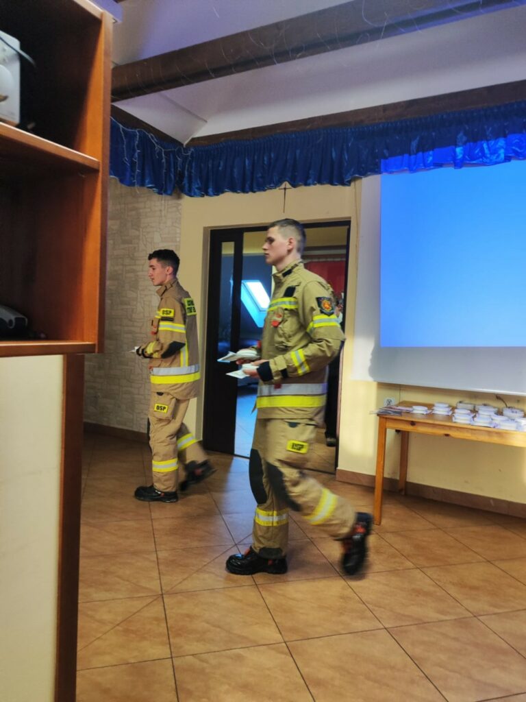 W pomieszczeniu przypominającym salę świetlicową lub konferencyjną dwóch strażaków w umundurowaniu OSP przechodzi obok stołu. Na stole znajdują się zapakowane czujniki dymu. Strażacy mają na sobie beżowe ubrania ochronne z elementami odblaskowymi i czarne buty. W tle widać ekran projekcyjny oraz drzwi prowadzące do innego pomieszczenia.