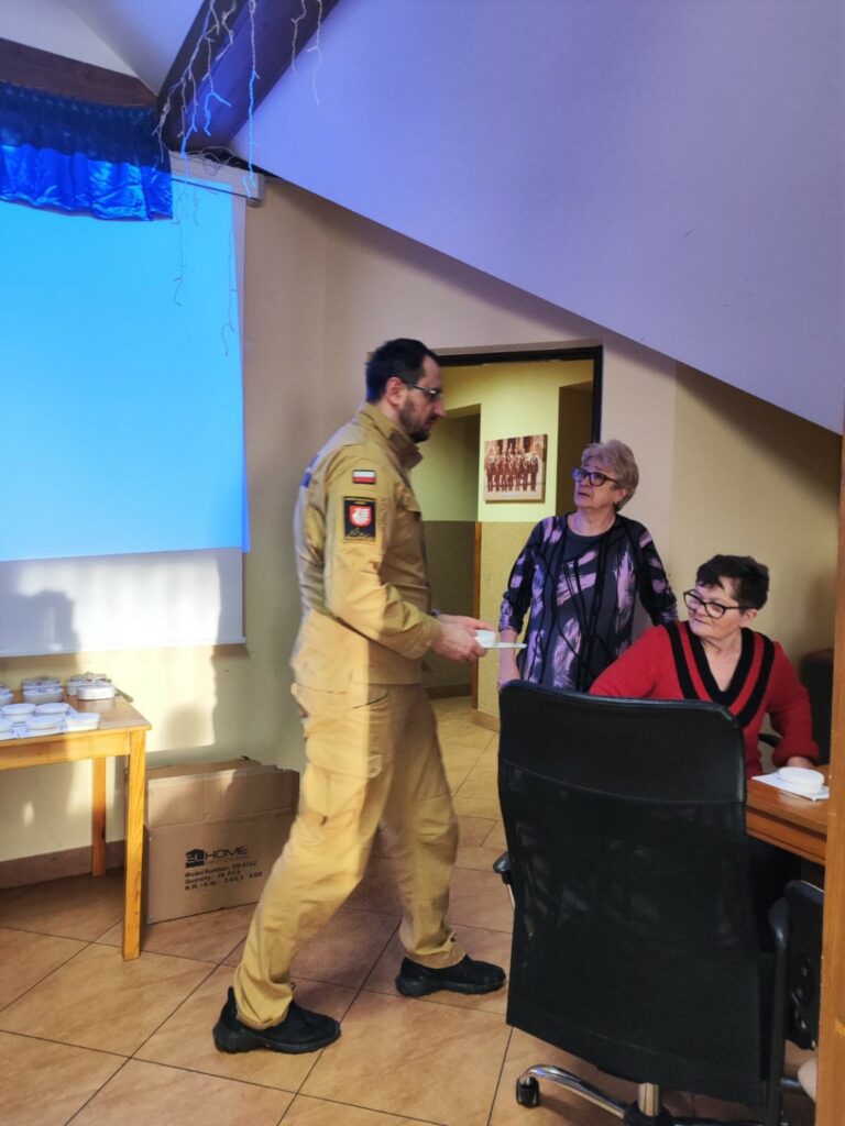 W sali spotkań strażak w umundurowaniu OSP wręcza niewielkie urządzenie kobiecie siedzącej przy stole. Obok niej stoi druga kobieta i obserwuje rozmowę. Na stoliku w tle leżą zapakowane czujniki dymu, a przy ścianie stoi karton z kolejnymi urządzeniami. W pomieszczeniu znajduje się ekran projekcyjny oraz dekoracyjne oświetlenie przy suficie.