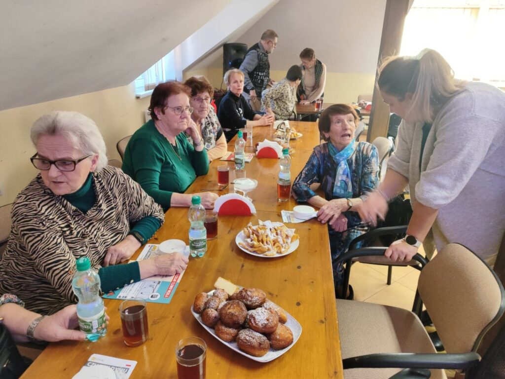 Przy długim drewnianym stole siedzi grupa starszych osób podczas spotkania. Na stole stoją szklanki z herbatą, butelki wody oraz talerze z pączkami i faworkami. Jedna z kobiet rozmawia z młodszą osobą stojącą obok, która pochyla się nad stołem. Część uczestników ma przed sobą materiały informacyjne oraz czujniki dymu. W tle widać kolejne osoby oraz sprzęt nagłaśniający.